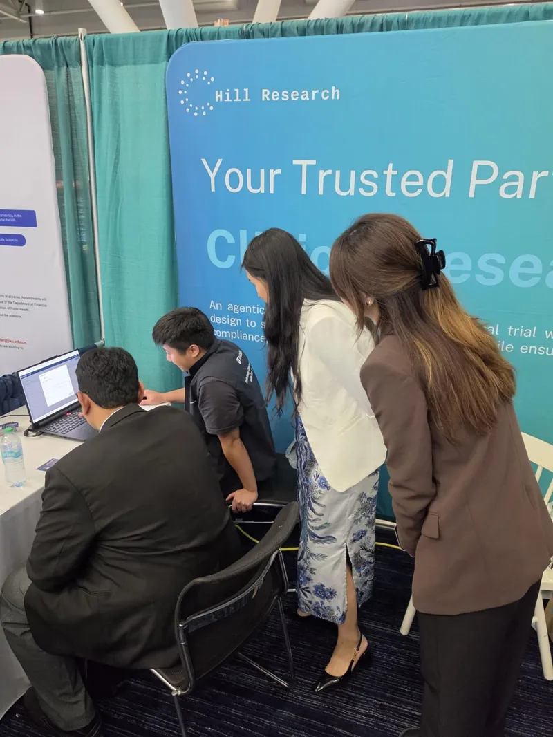 Visitors gathered around a laptop at Hill Research's Booth 607 in the EXPO hall at JSM 2026, in front of the Hill Research backdrop